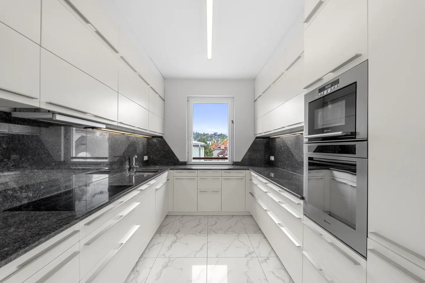 A modern kitchen with white cabinets, black granite countertops, and marble floors. A window overlooks a cityscape.