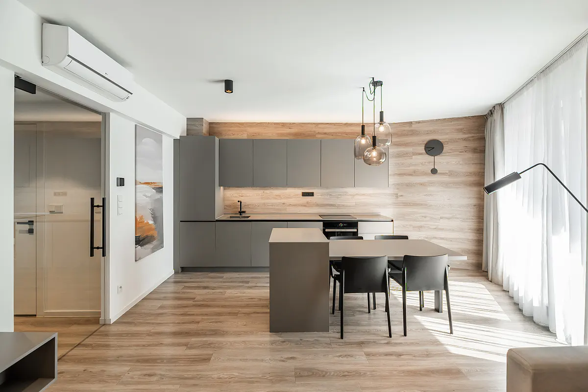 Modern kitchen with grey cabinets, wood-look flooring, a dining table with black chairs, and large windows with sheer curtains.
