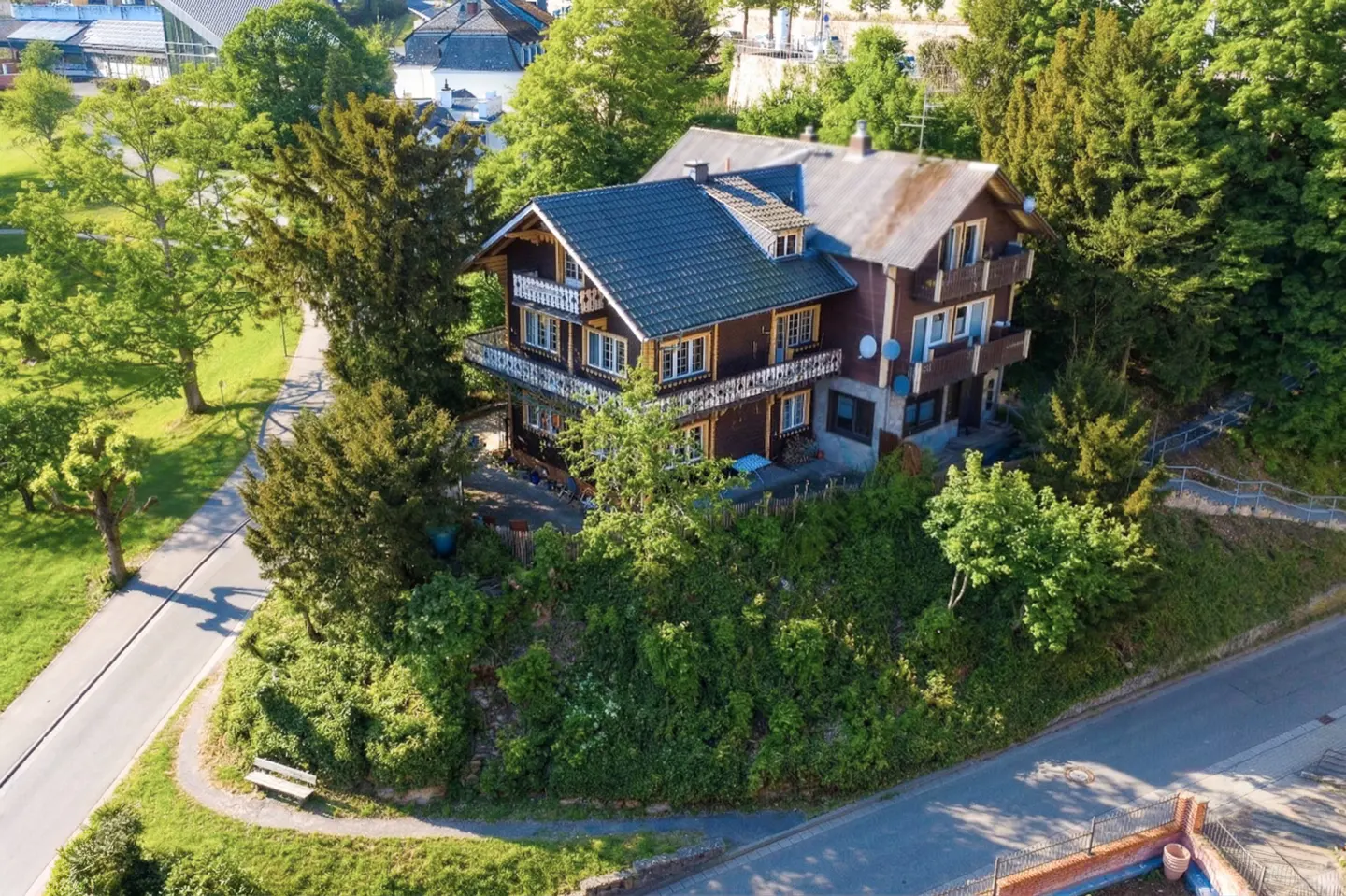 A brown, multi-story house with a blue roof sits on a green, tree-covered hill. A road and bench are visible in the foreground.