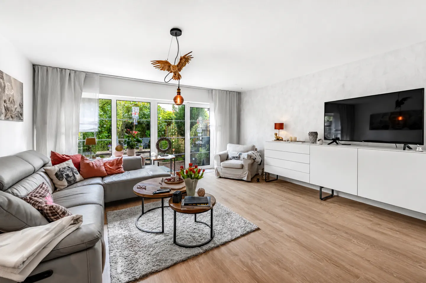 Bright living room with gray sectional, wood floors, and white walls. A unique bird light fixture hangs above round coffee tables. Large windows overlook a green yard.