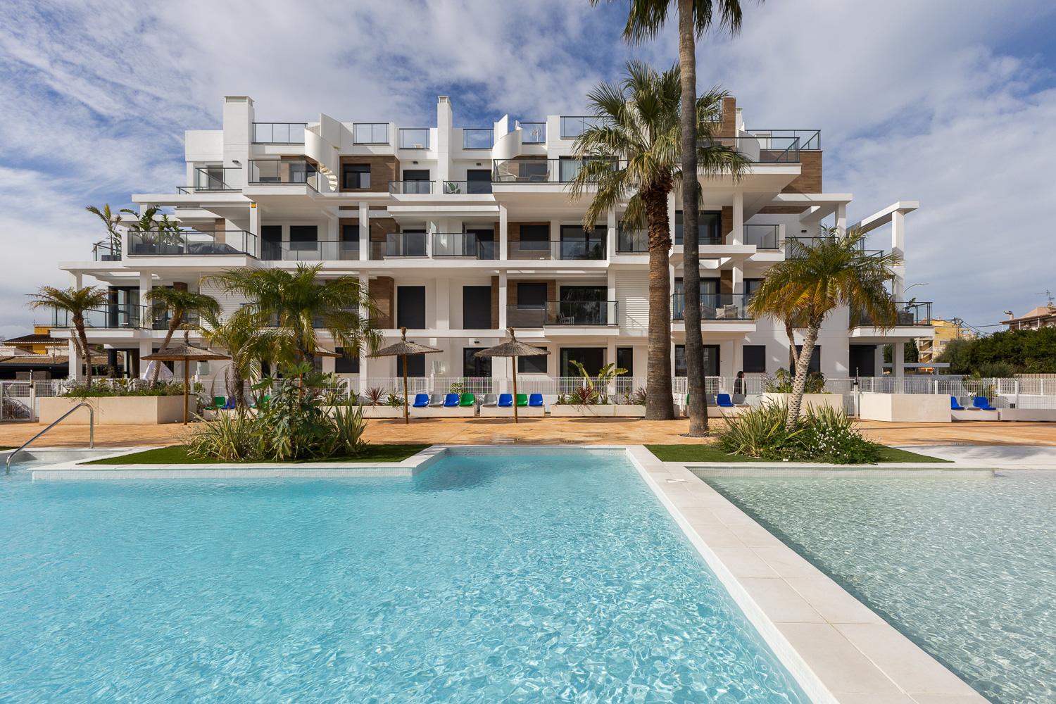 Modern white apartment building with balconies, palm trees, and a turquoise swimming pool in the foreground.