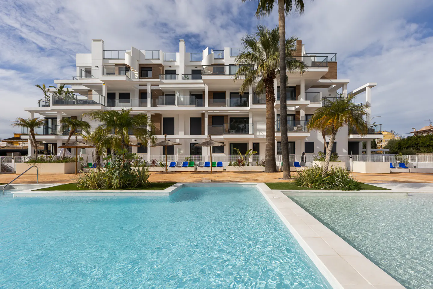 Modern white apartment building with balconies, palm trees, and a turquoise swimming pool in the foreground.
