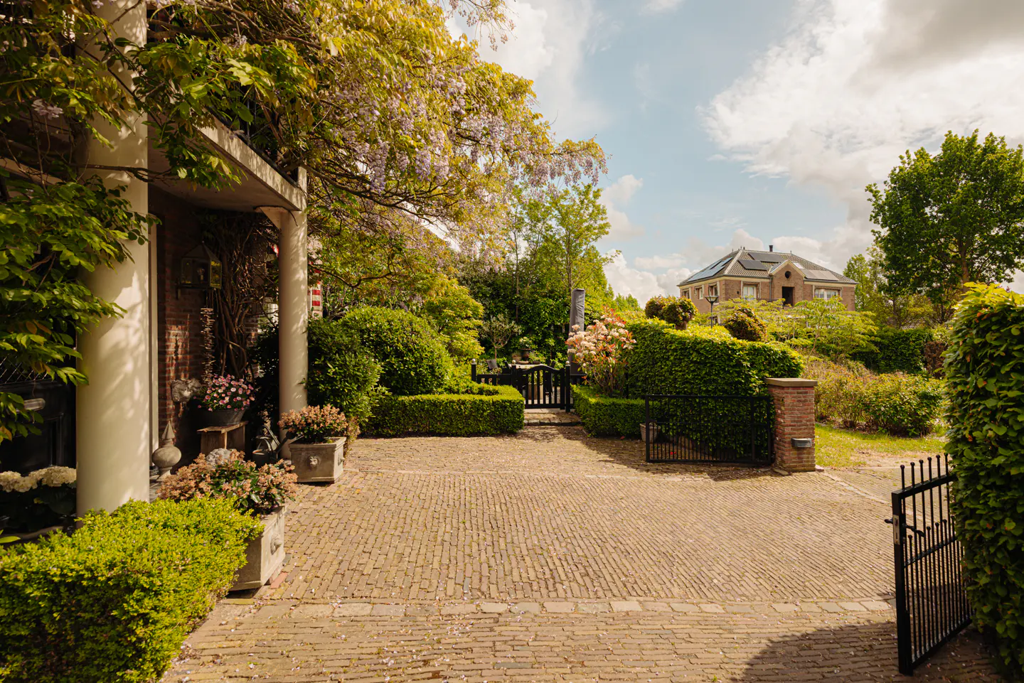 Brick driveway leads to a home with lush landscaping and a black metal gate. The sky is blue with white clouds.