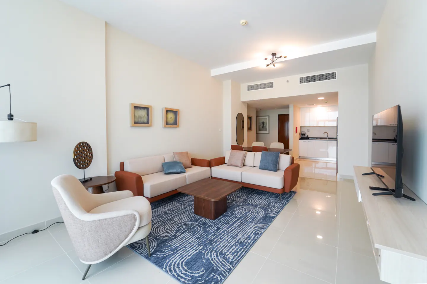 Bright living room with cream sofas, a blue patterned rug, and a dark wood coffee table. A kitchen is visible in the background.