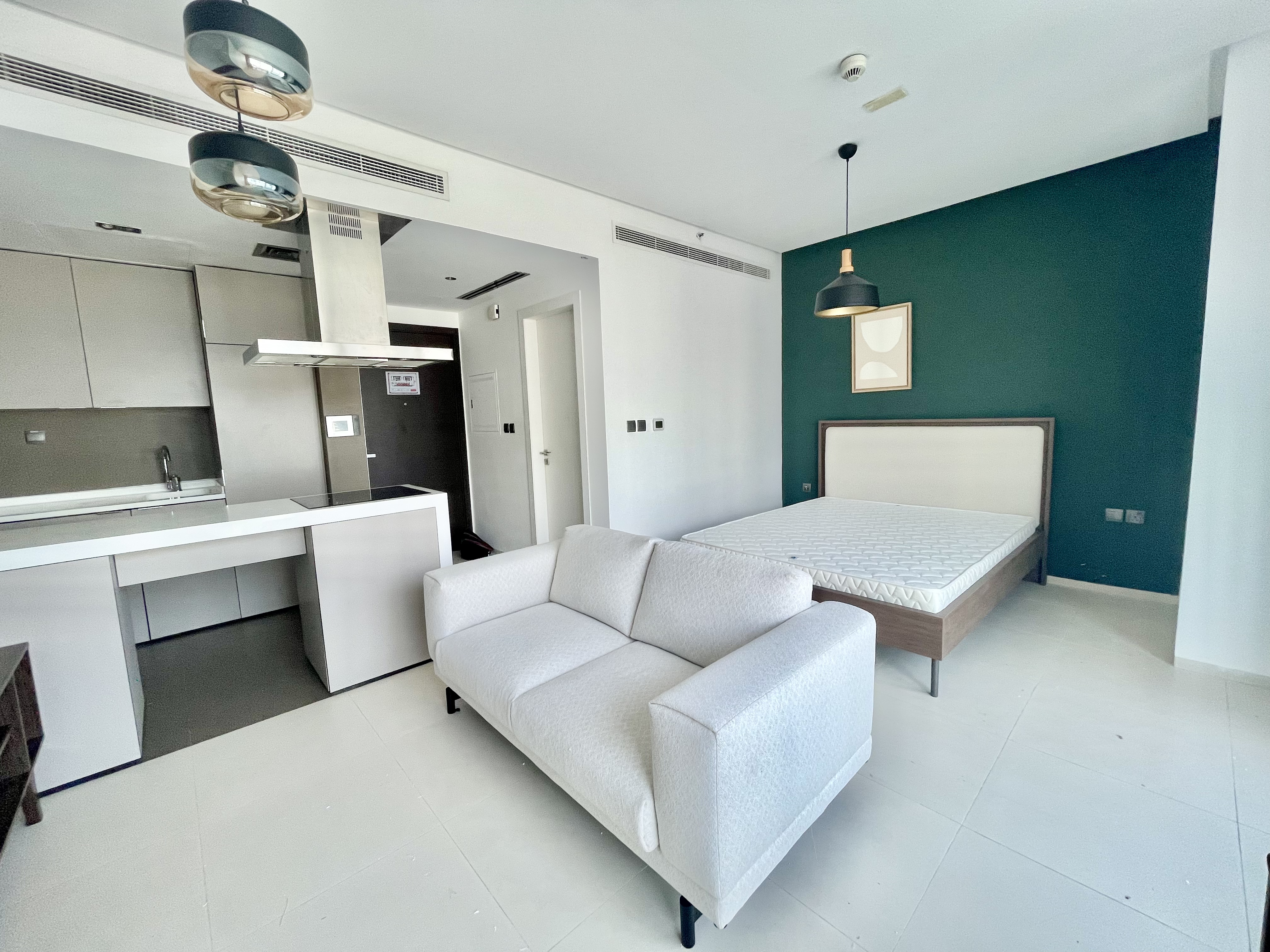 Modern studio apartment with a white sofa, a bed against a dark green wall, and a sleek kitchen with light gray cabinets.