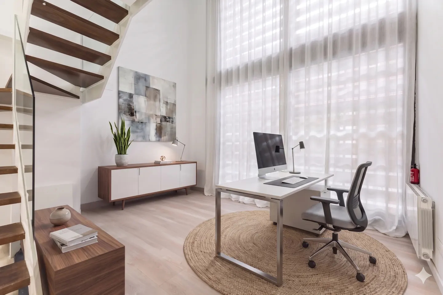 Bright home office with white walls, wood stairs, and large windows with sheer curtains. A white desk, computer, and gray chair sit on a round jute rug.
