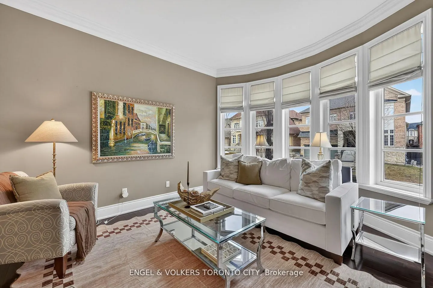 Bright living room with a white sofa, patterned armchair, glass coffee table, and a painting of a canal on a beige wall.