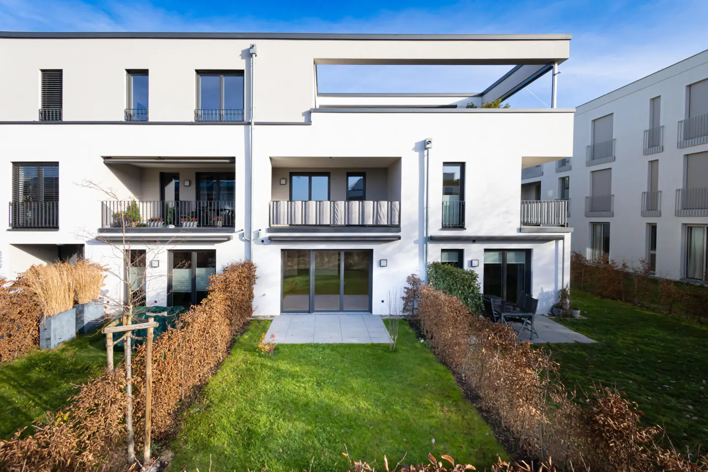 Modern white townhouses with balconies and patios overlook a green lawn and trimmed hedges under a bright blue sky.