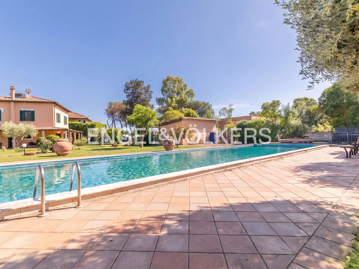 Outdoor pool with blue water and a brick patio, surrounded by green grass, trees, and a peach-colored house under a clear blue sky.