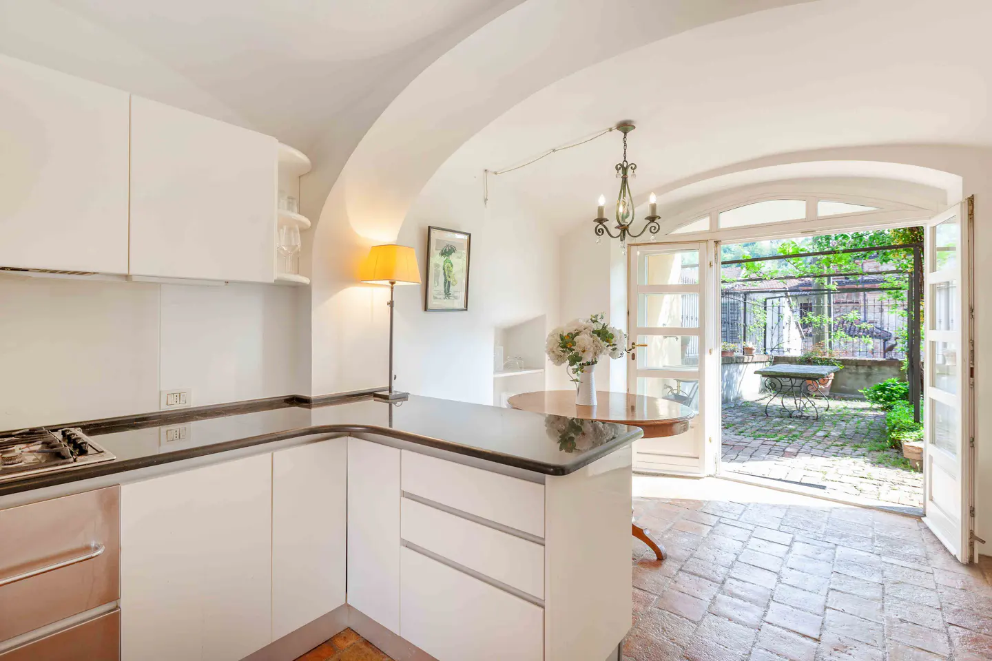 Bright kitchen with white cabinets, black countertops, and brick floors. Open doors lead to a lush green courtyard. A round table with flowers sits nearby.