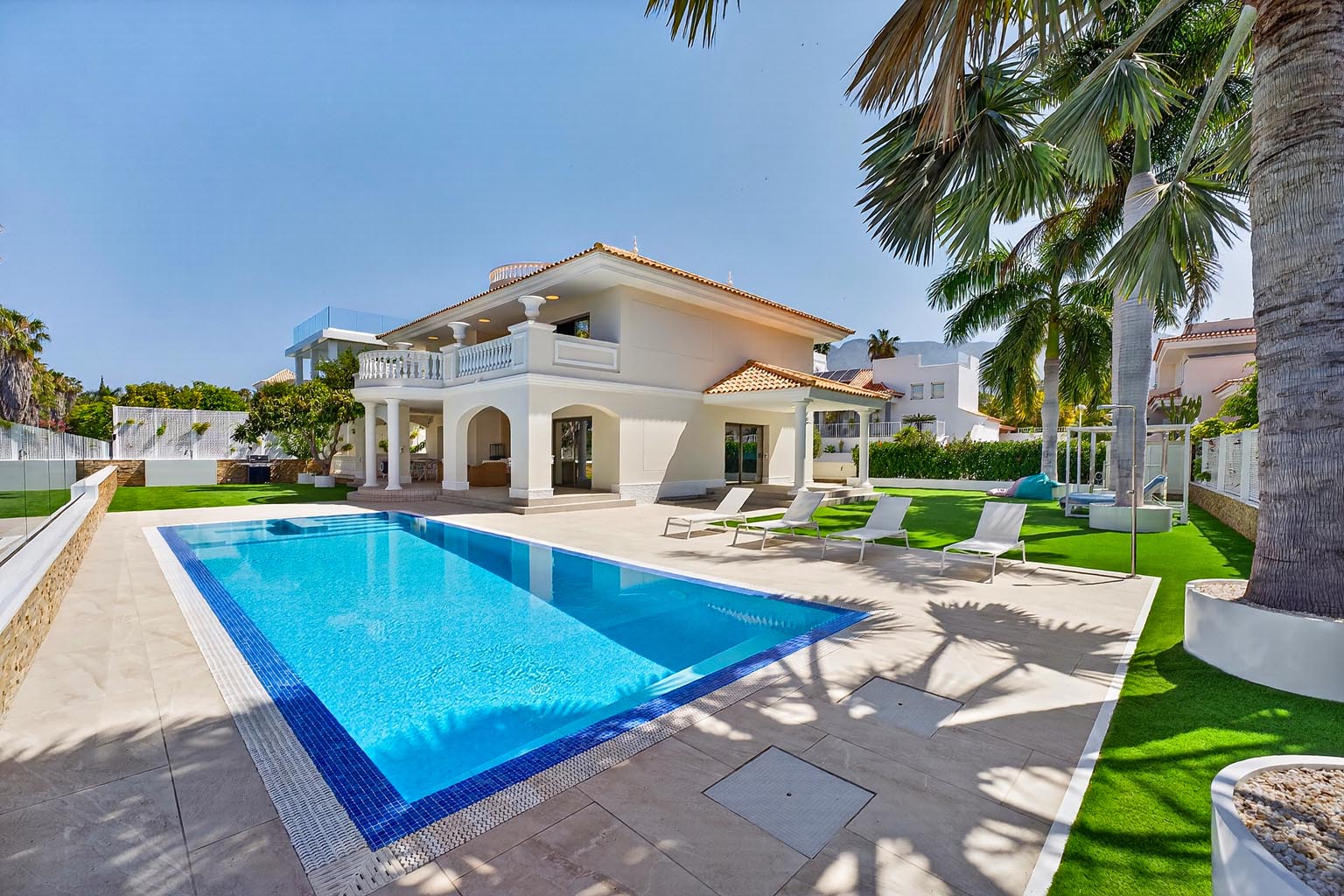 Exterior view of a two-story white house with a blue-tiled pool, lounge chairs, and palm trees on a sunny day.