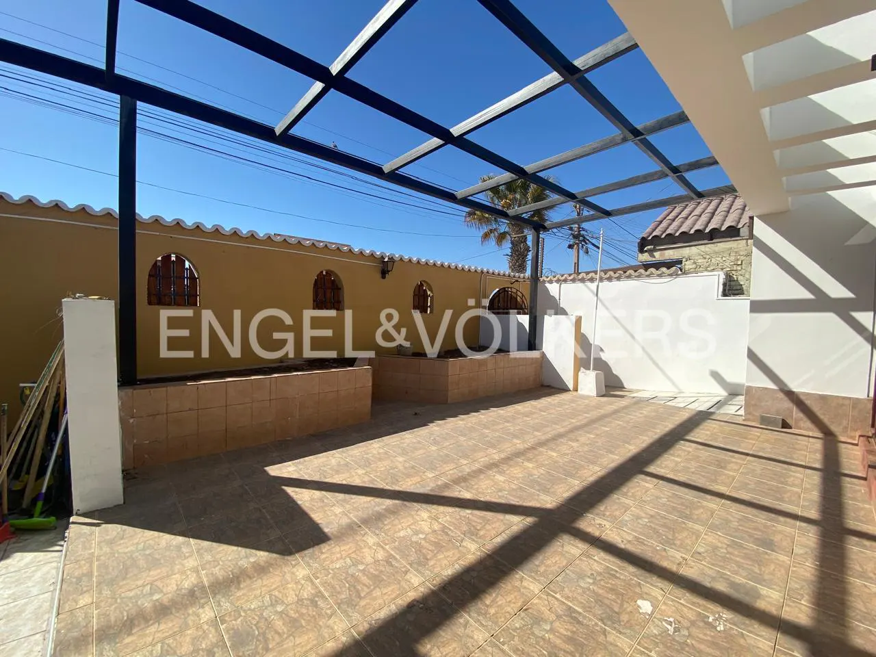 Outdoor patio with tan tile flooring, a black metal pergola, and a yellow wall with arched windows. The Engel & Völkers logo is superimposed.
