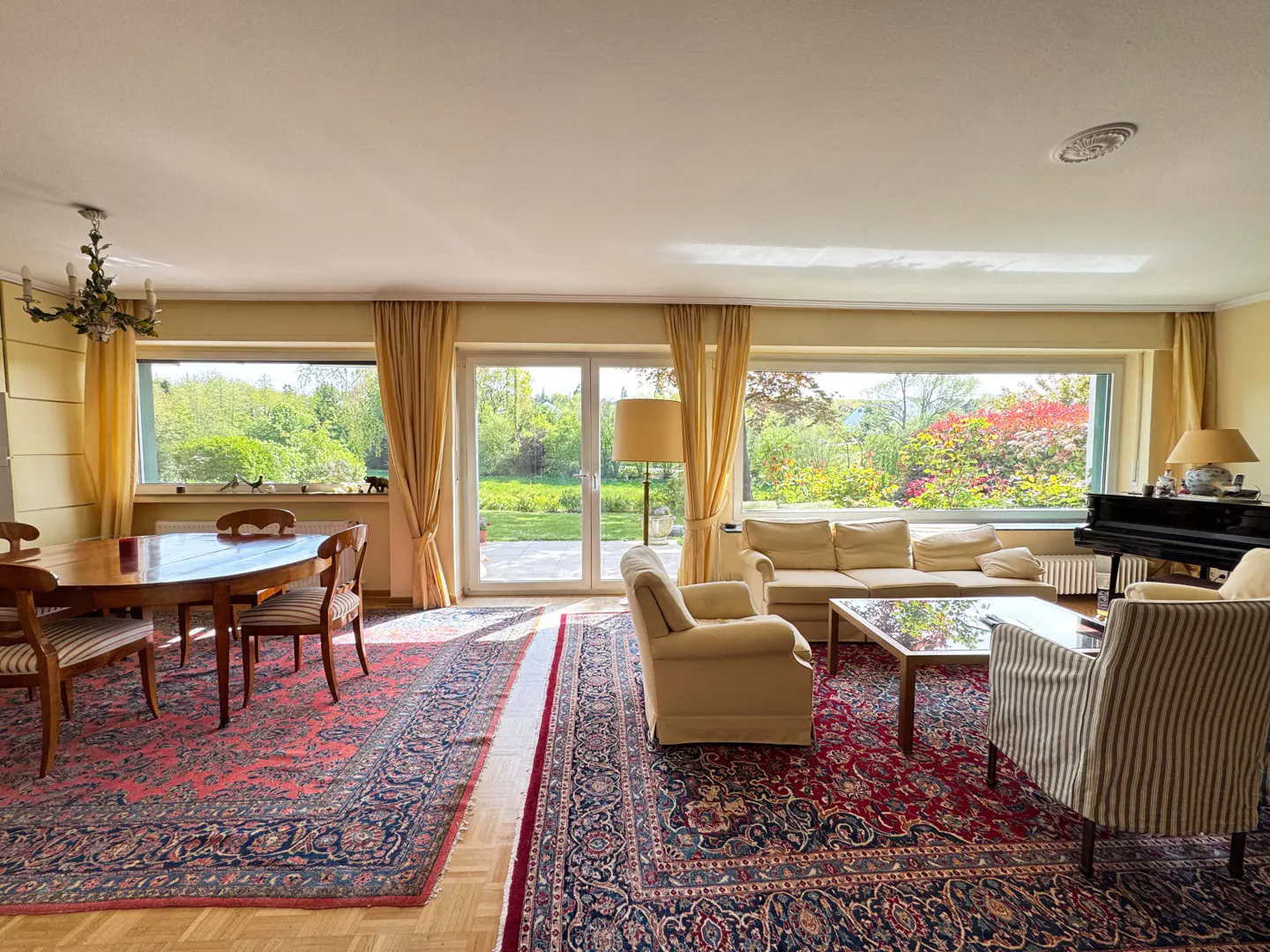 Bright living room with dining table, sofas, armchairs, piano, and large windows overlooking a green garden. Red patterned rugs on wood floors.