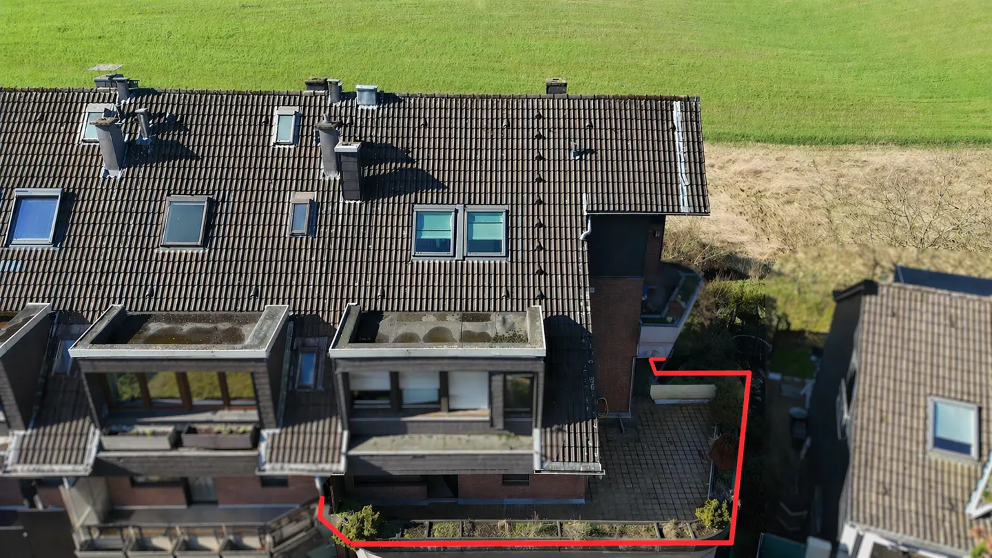 Aerial view of a brown-roofed townhouse with dormer windows, chimneys, and a red-outlined patio, set against a green field.