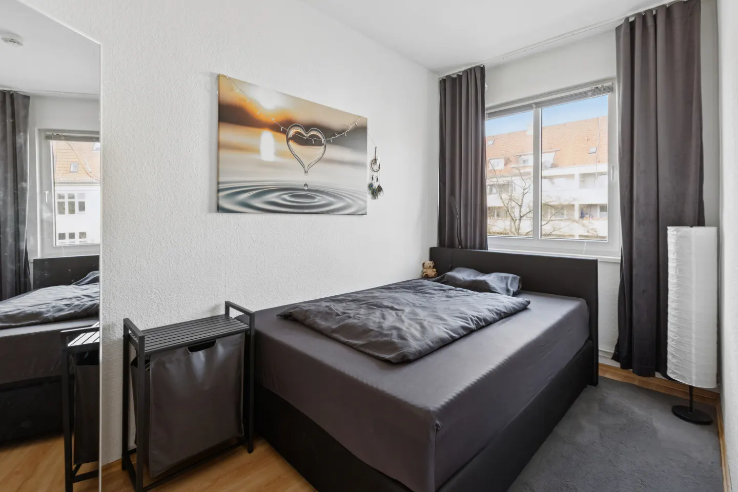 A bedroom with a gray bed, gray curtains, and a picture of a heart over water. A laundry basket is next to the bed.