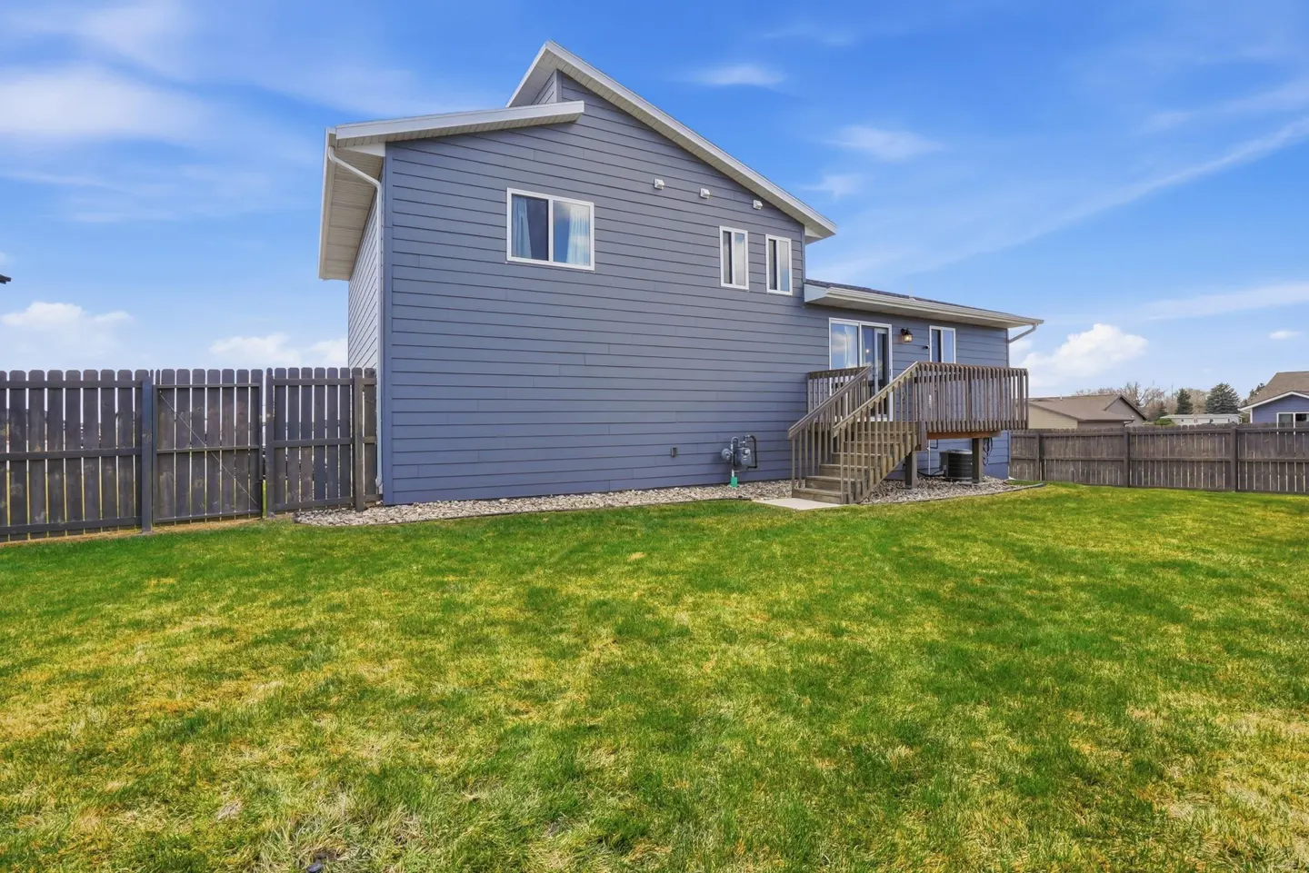 Backyard view of a two-story gray house with a wooden deck and stairs, surrounded by a green lawn and a dark gray wooden fence.