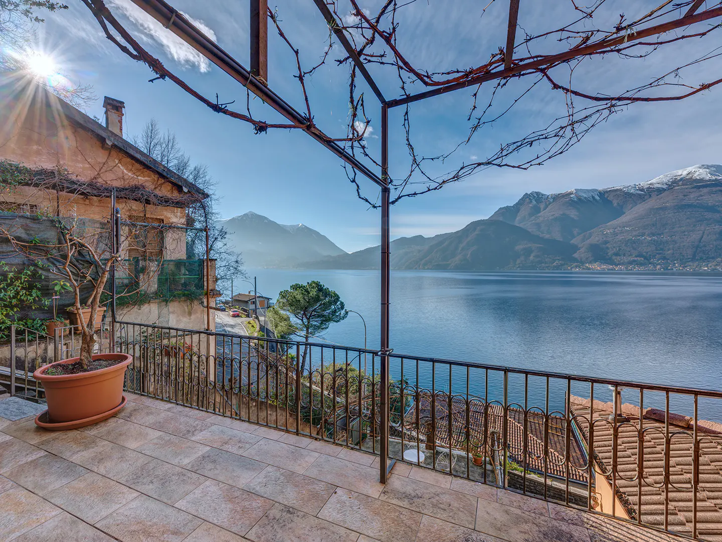 Balcony view of Lake Como, Italy. Mountains in the background, blue water, and a metal pergola frame with vines overhead.