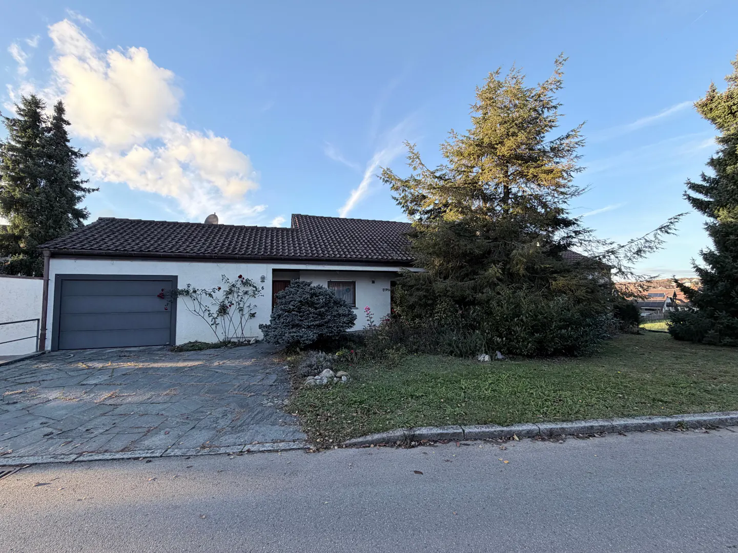 A single-story white house with a gray garage door and a brown tiled roof, surrounded by green trees and grass.