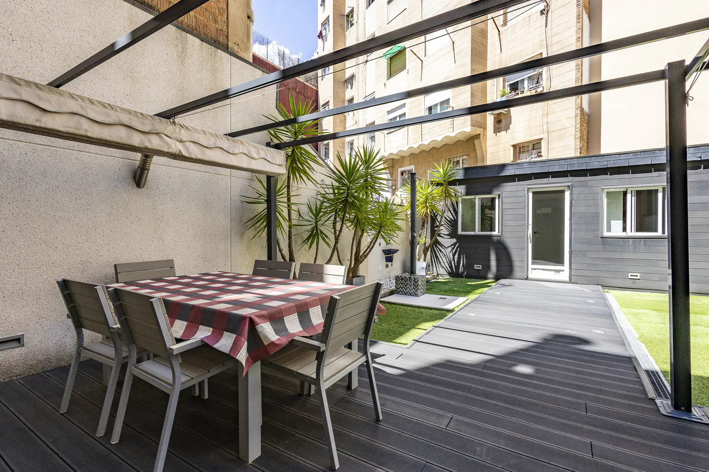 Outdoor patio with a table and chairs under a metal pergola, with a small gray building and green plants in the background.