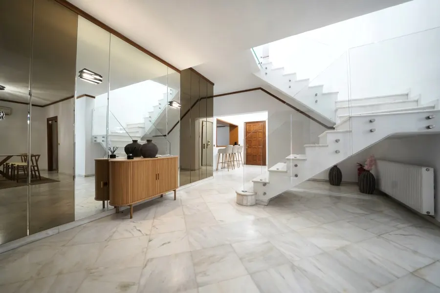 Bright foyer with marble floors, mirrored wall, and white staircase with glass railing. A wooden cabinet sits against the mirrored wall.