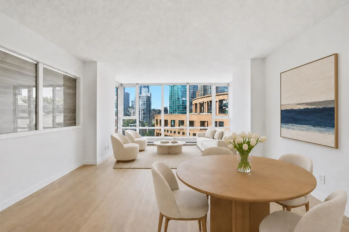 Bright, modern apartment with city views. A round wood table with white chairs sits near a vase of white tulips. Living area with white sofa and chairs.
