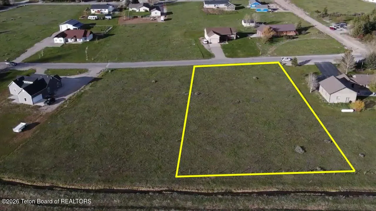 Aerial view of a vacant, grassy residential lot outlined in yellow, situated in a neighborhood with nearby houses.