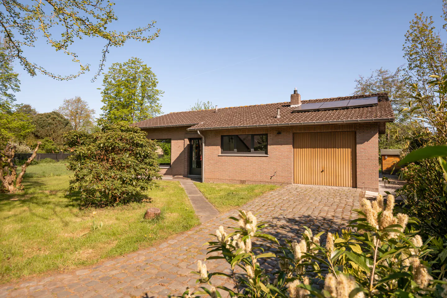 Exterior view of a single-story brick house with a brown roof, wooden garage door, and solar panels. A stone path leads to the entrance, surrounded by green lawn and trees.