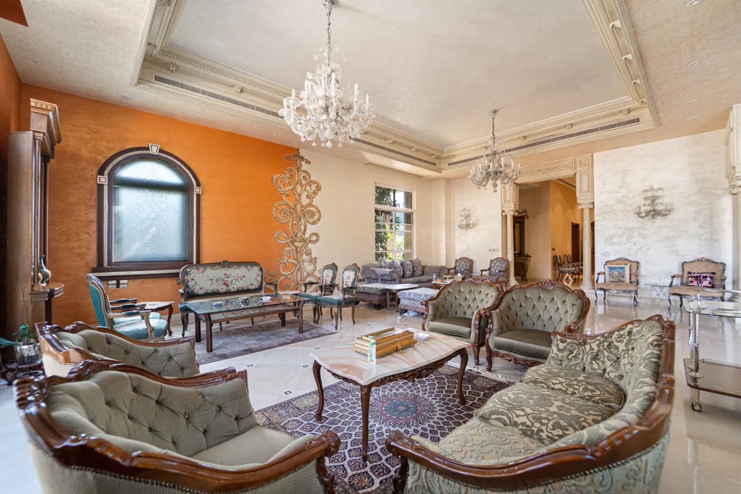 A luxurious living room with ornate sofas, chandeliers, and an orange accent wall.