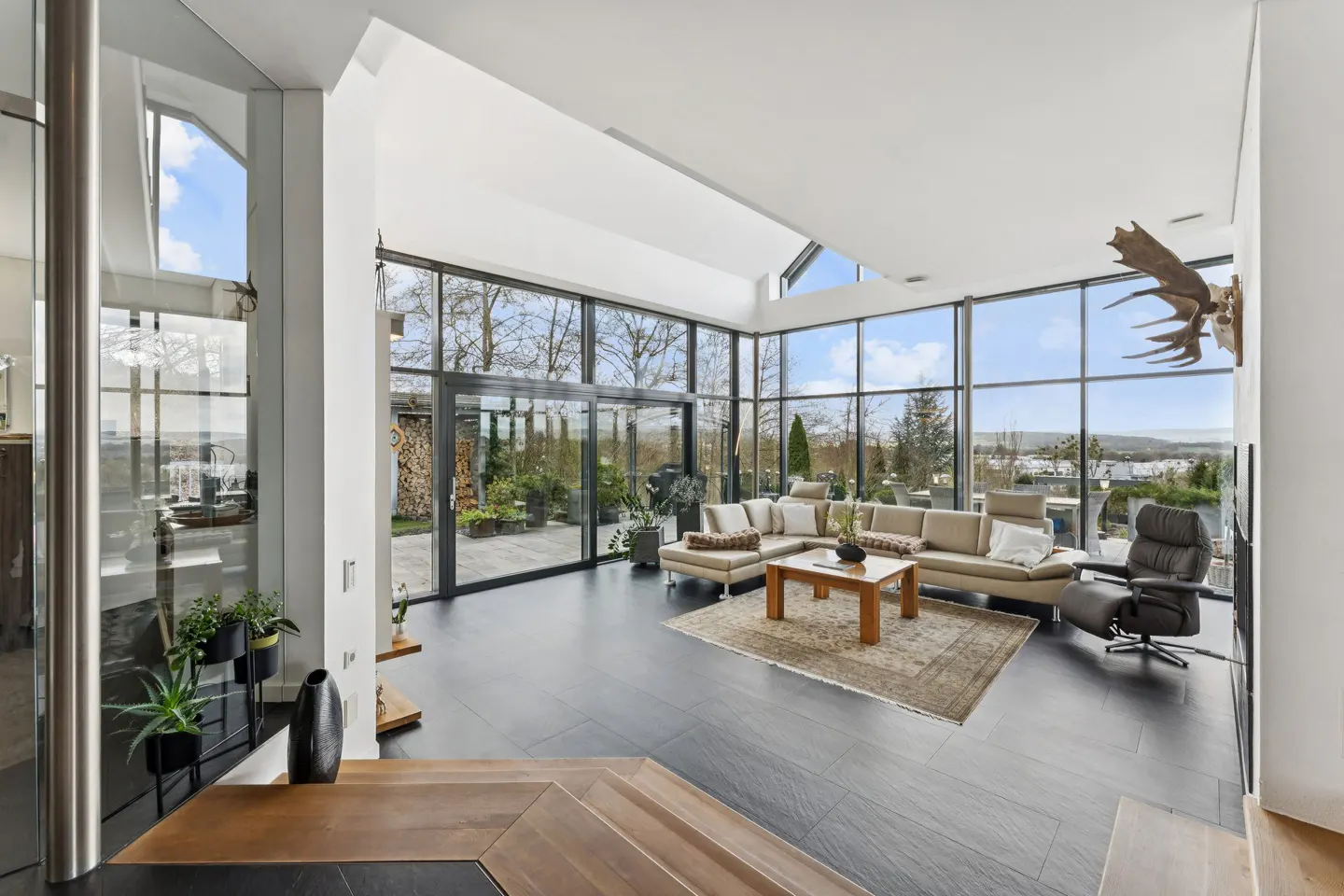 Bright living room with black tile floor, beige sectional sofa, wood coffee table, and floor-to-ceiling windows with a view of trees.