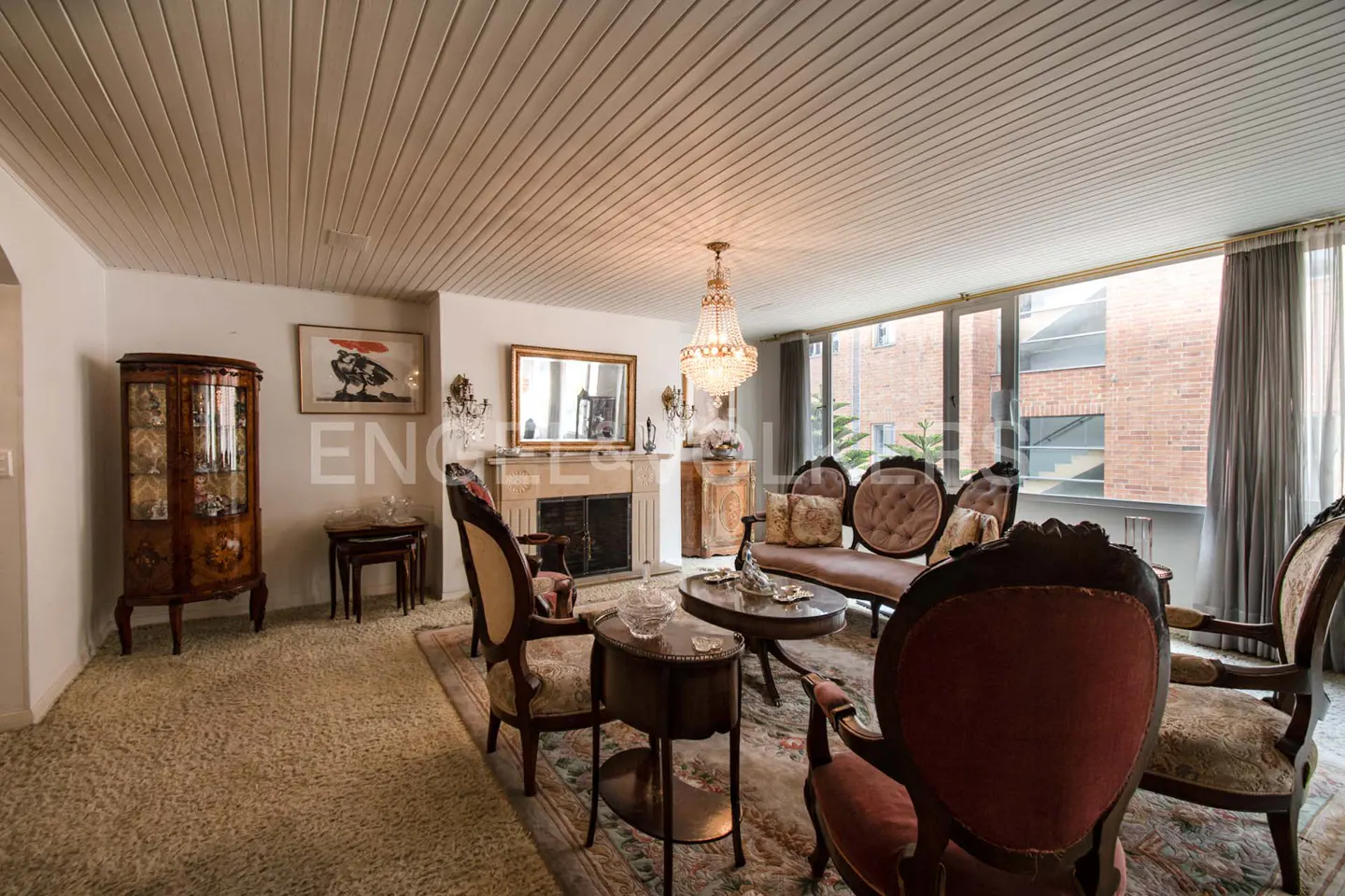 Living room with beige carpet, white walls, and a wood-paneled ceiling. Antique furniture includes a china cabinet, chairs, and a pink sofa. A chandelier hangs above.