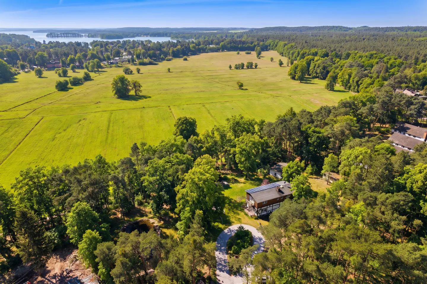 Aerial view of a property with a large green field, trees, and a lake in the background under a blue sky.