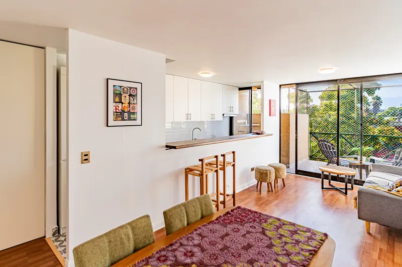 Bright apartment interior with wood floors, white walls, and a balcony view of lush greenery. Dining table with floral runner in foreground.