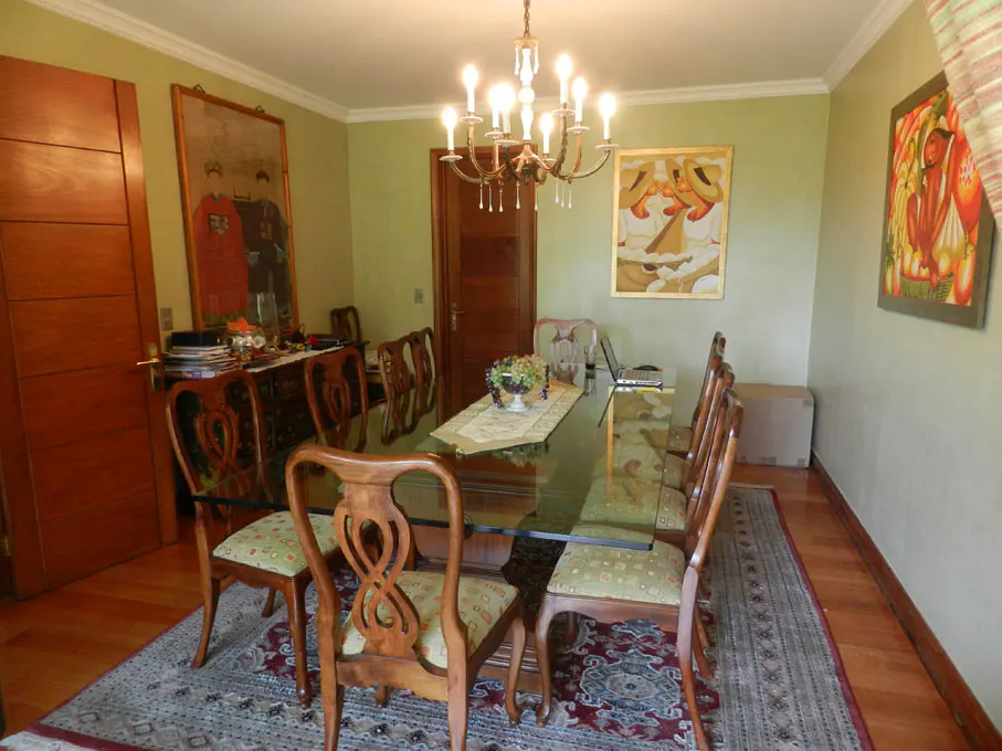 Dining room with a glass-top table, wooden chairs, and a chandelier. Paintings adorn the light green walls. A patterned rug covers the wooden floor.