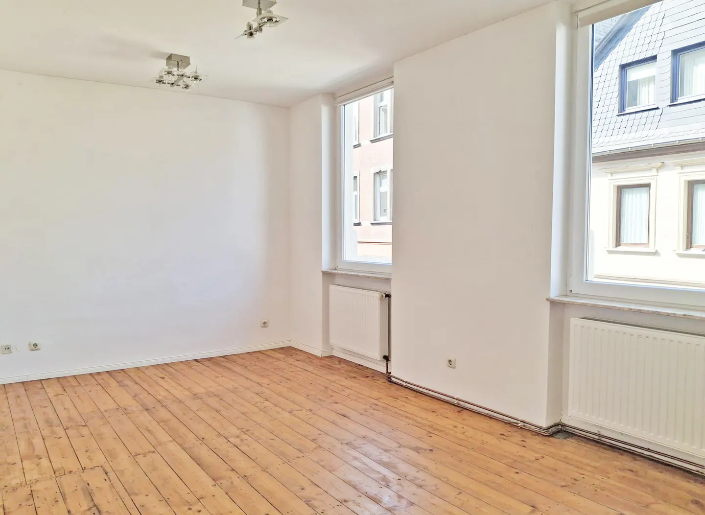 Bright, empty room with hardwood floors, white walls, two windows, and two ceiling lights.