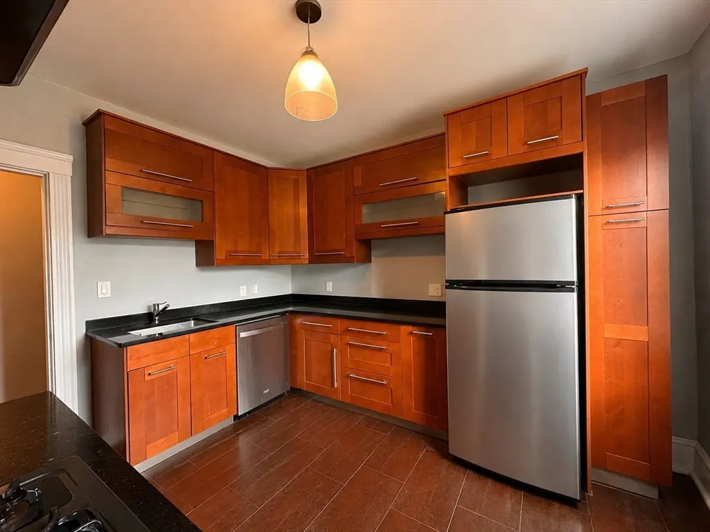A kitchen with wood cabinets, stainless steel appliances, black countertops, and brown tile floors.