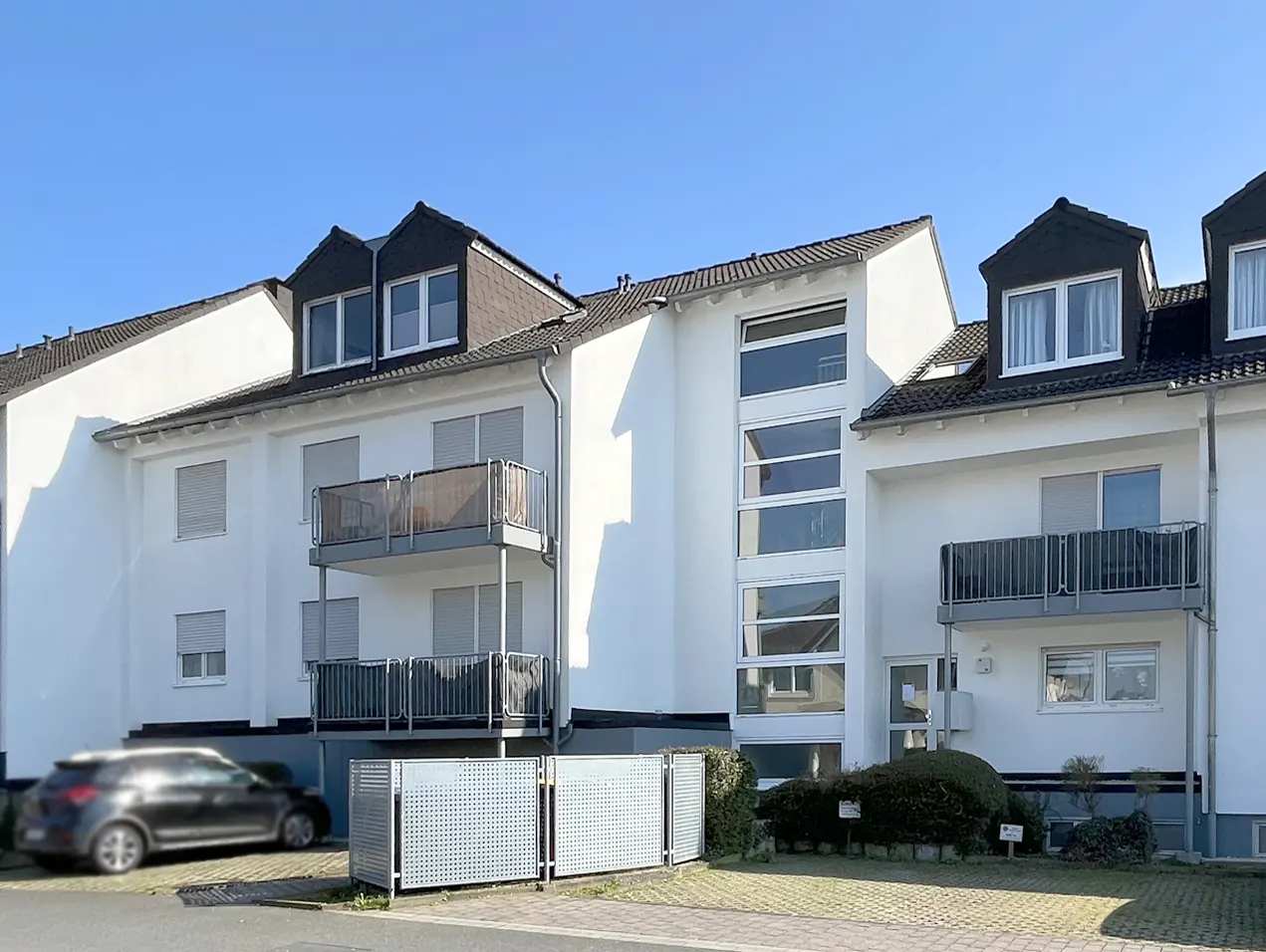 Exterior of a modern, white apartment building with balconies and dark roof tiles under a clear blue sky. A car is parked nearby.