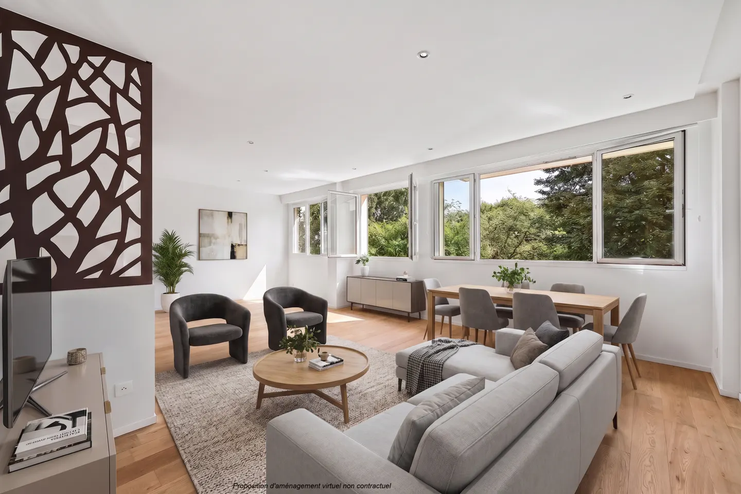 Bright living room with hardwood floors, a gray sofa, two chairs, and a dining table with a view of green trees through the windows.