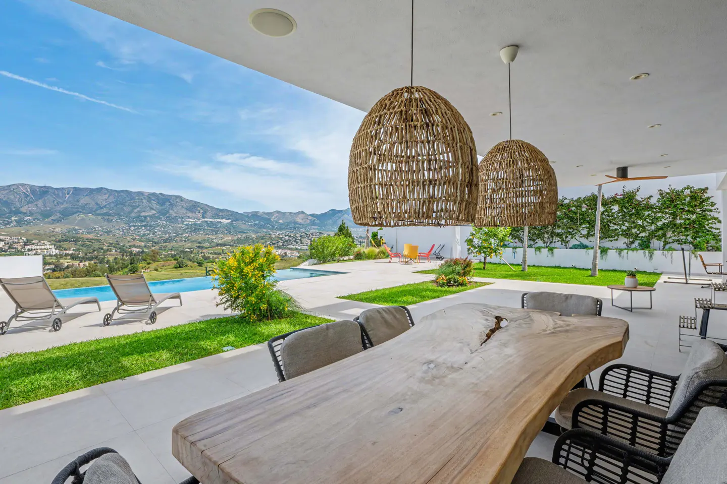 Outdoor patio with a large wooden table, wicker pendant lights, and a view of a pool, mountains, and blue sky.