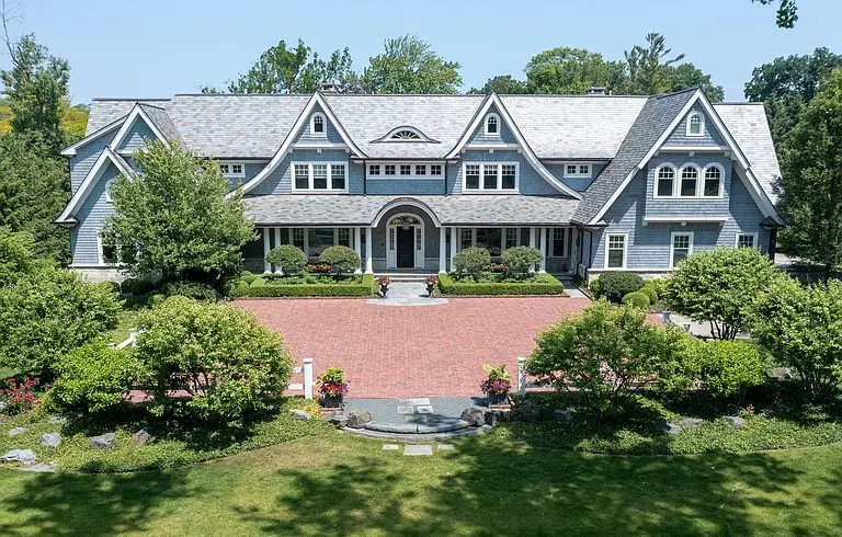 Large, two-story blue house with a gray roof and a red brick driveway, surrounded by green trees and lawn.