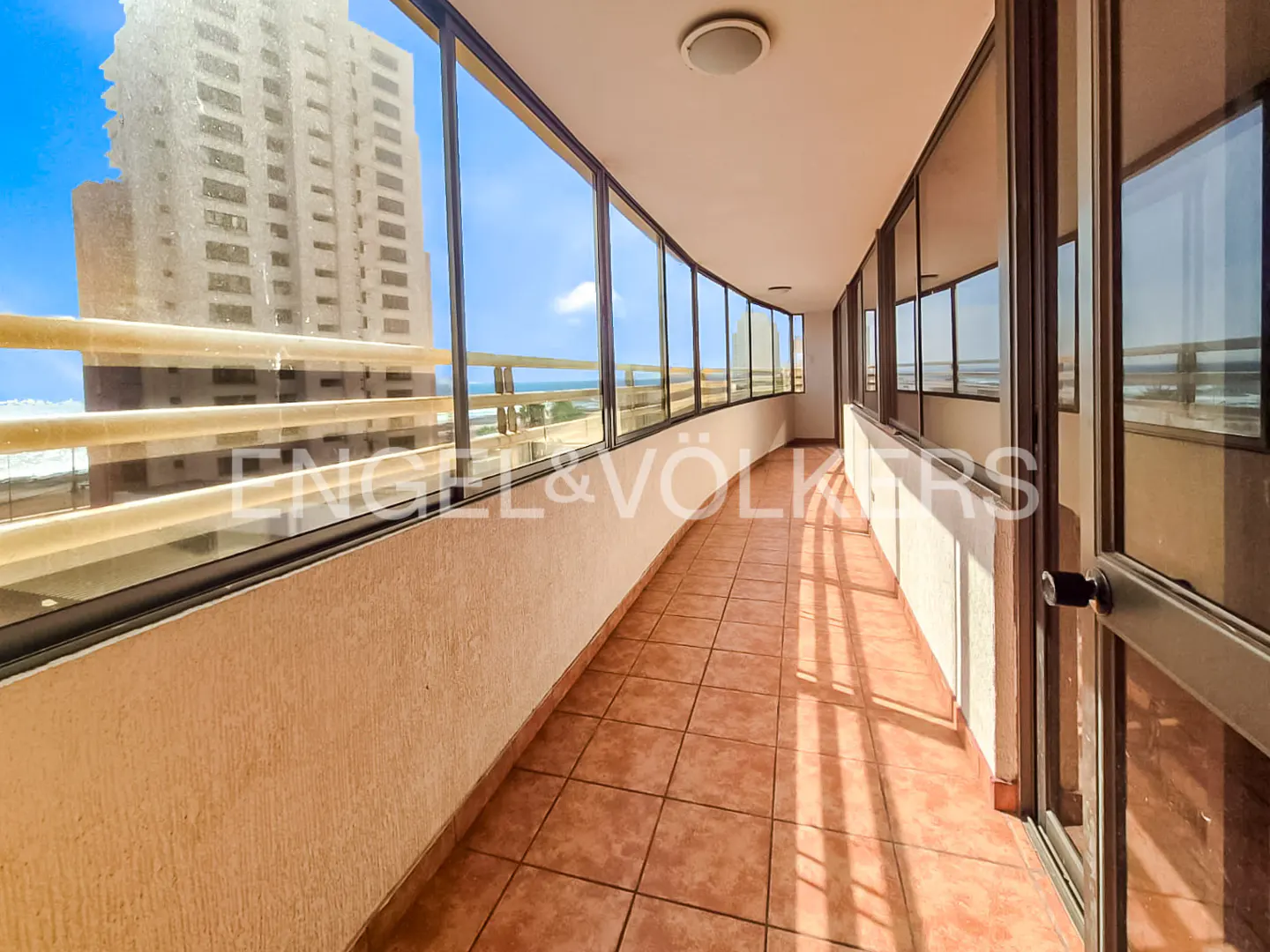Enclosed balcony with tile floor, floor-to-ceiling windows, and ocean view. Tall building visible through windows.