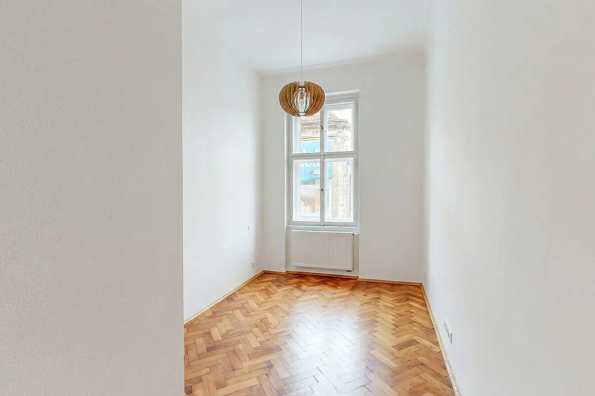 Empty room with white walls, herringbone wood floor, and a window with a white frame. A modern light fixture hangs from the ceiling.