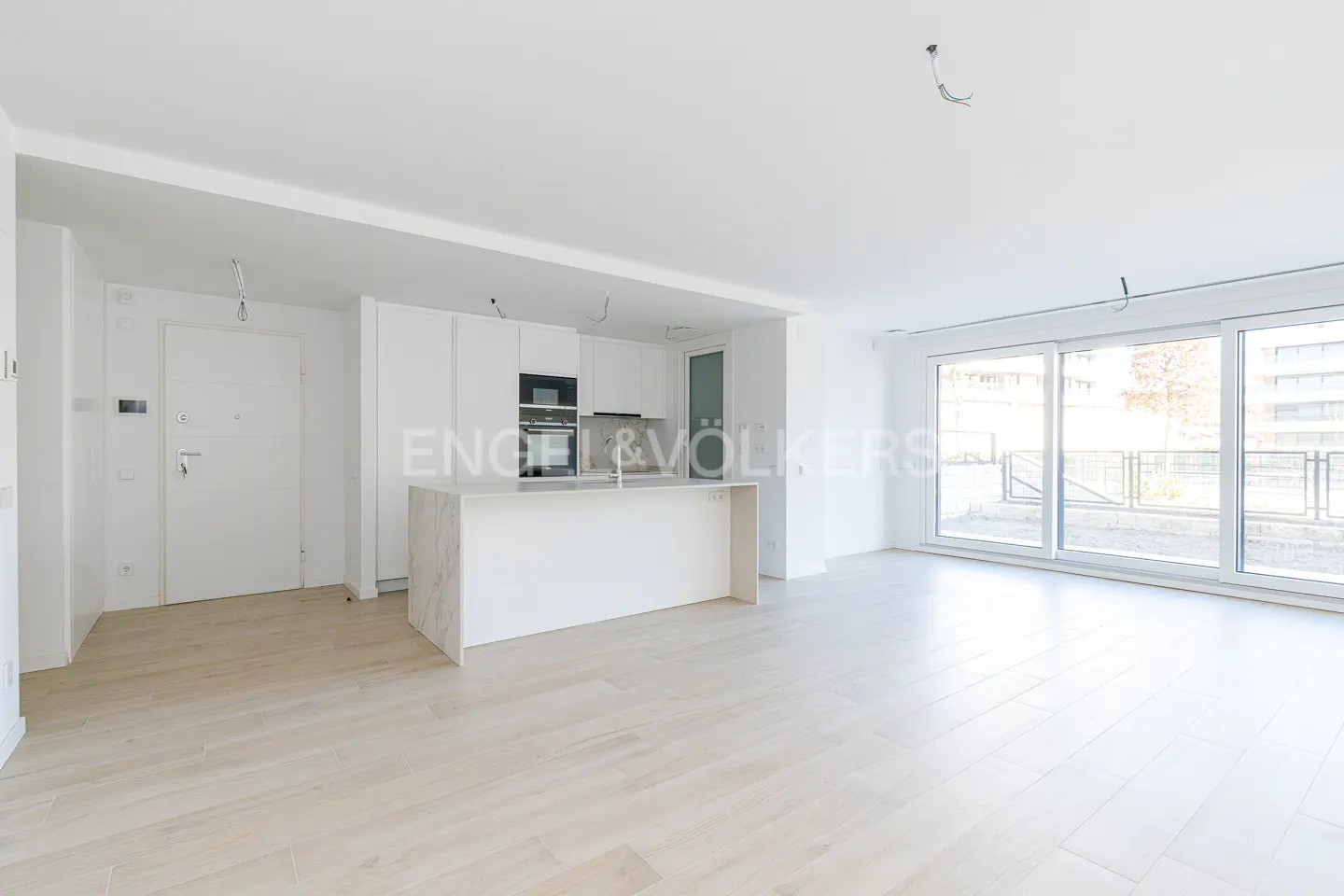 Bright, modern apartment with white walls, light wood floors, and a kitchen island. Large windows offer natural light.