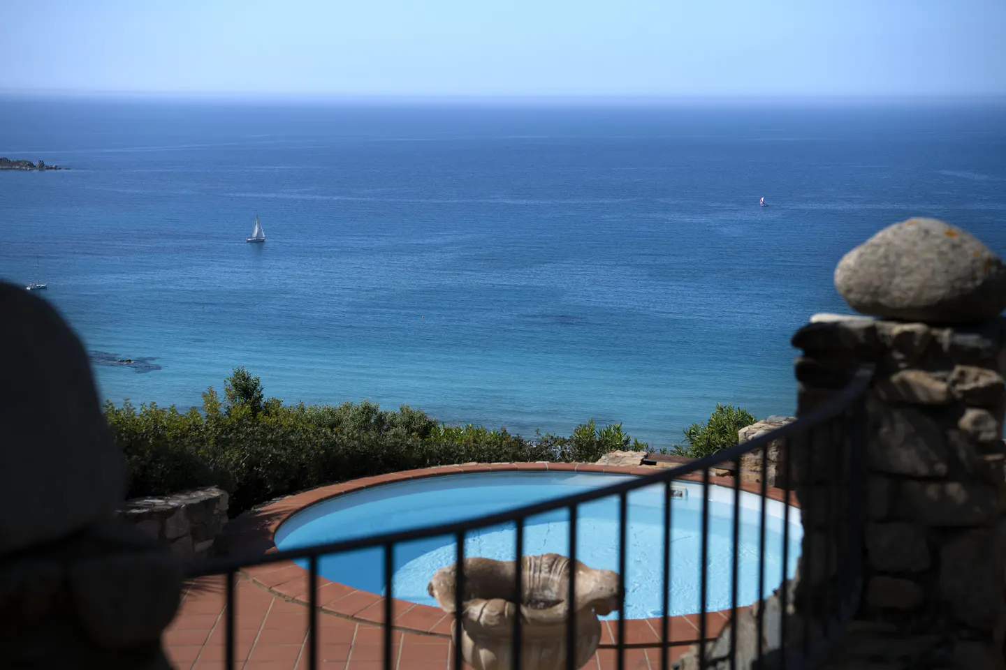 View of a blue pool overlooking the ocean, with a sailboat in the distance. Stone wall and black railing in the foreground.