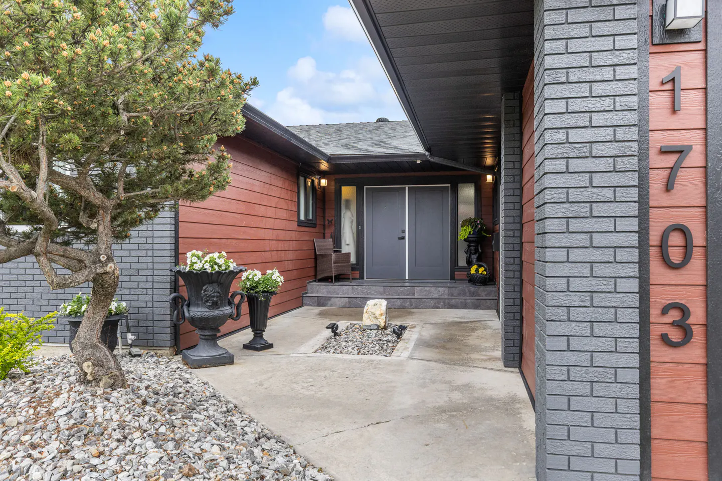 Exterior of a two-story house with red siding, gray brick, and a gray double door. The house number "1703" is visible on the right side. A tree and flower pots decorate the front.