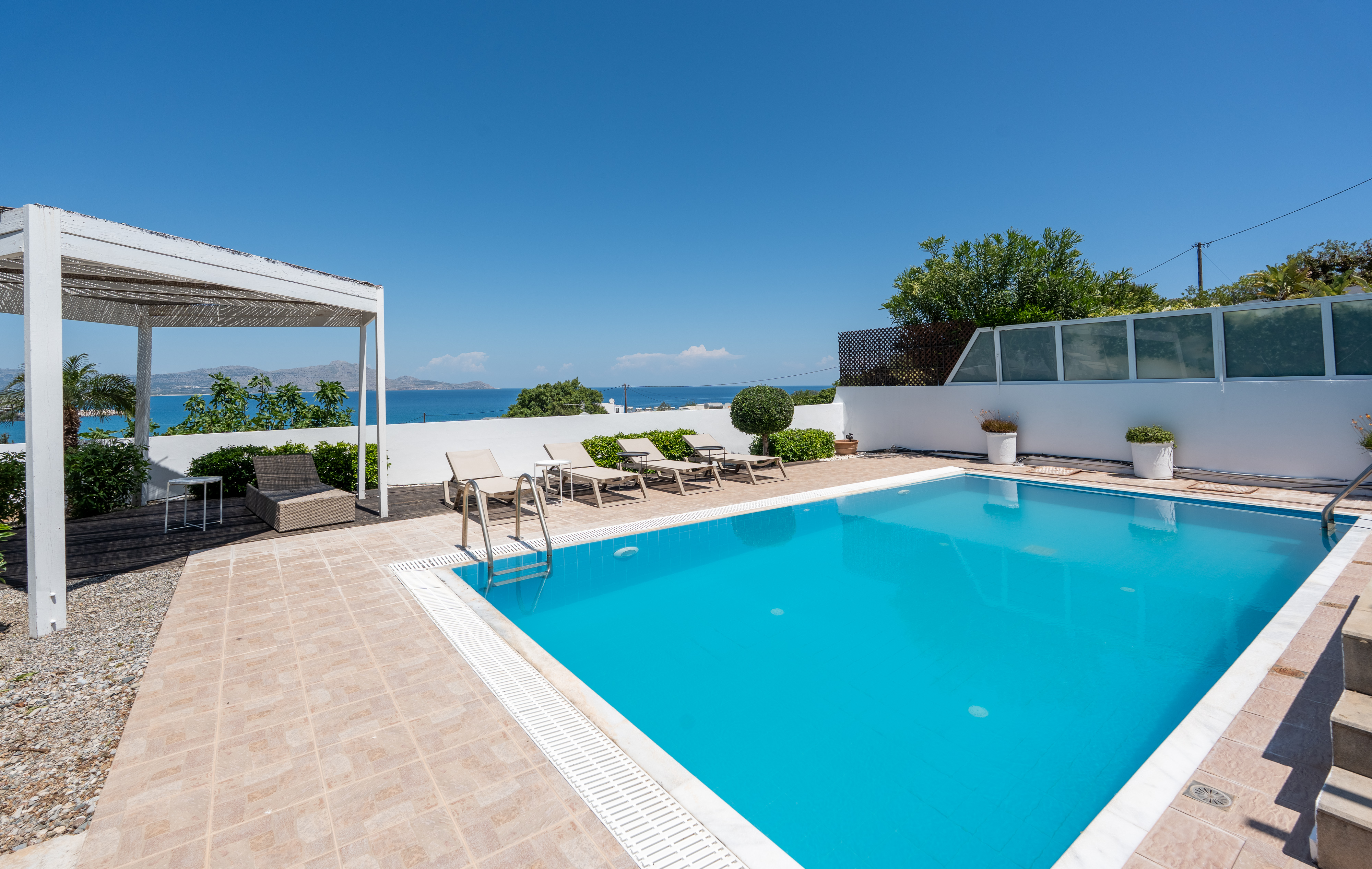 Outdoor pool area with blue water, lounge chairs, and a white pergola against a clear blue sky.