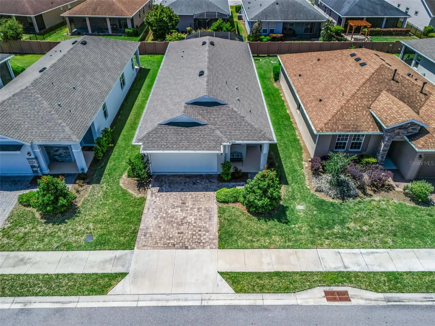 Aerial view of a single-story home with a gray roof, white garage door, and brick driveway in a suburban neighborhood.