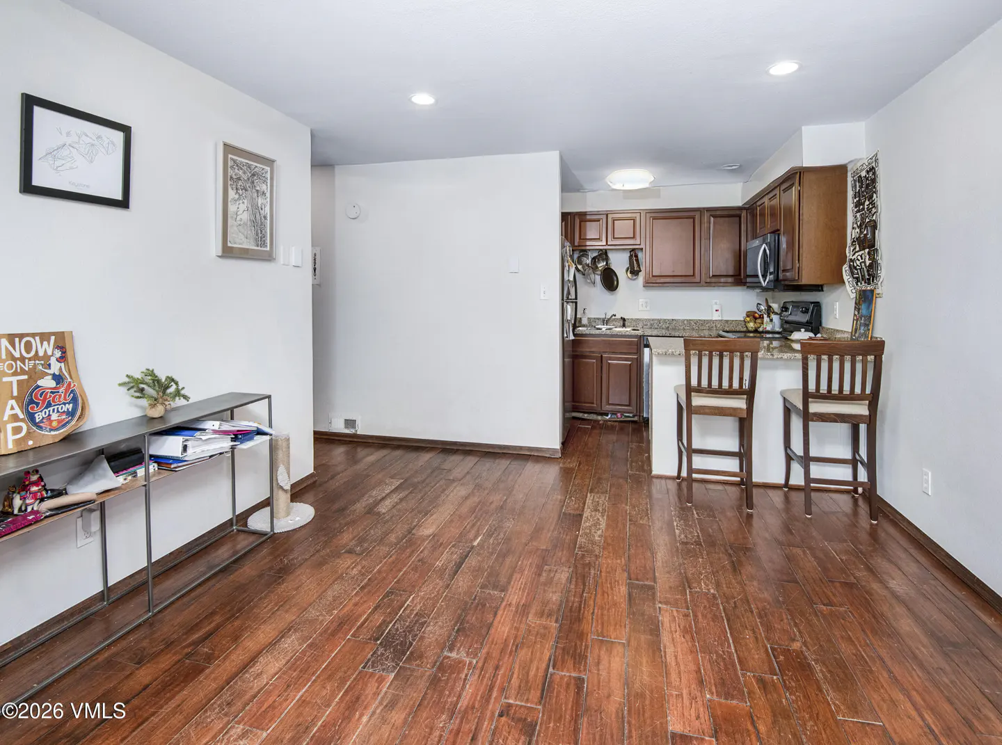 Open kitchen with dark wood cabinets, breakfast bar with two stools, and hardwood floors in a white-walled living space.