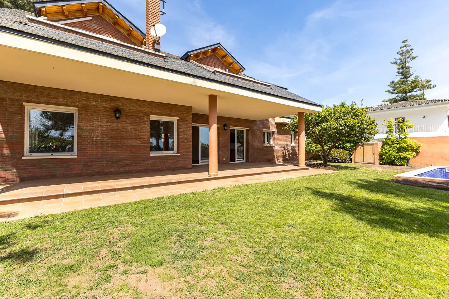 Brick house with a covered patio, supported by columns, overlooking a green lawn and a pool in the background.