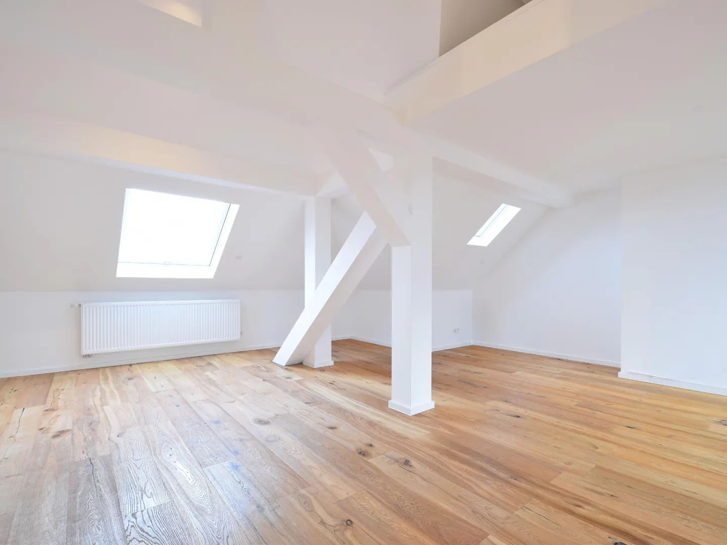 Bright, empty attic room with light wood floors, white walls, and white support beams. Two skylights and a radiator are visible.