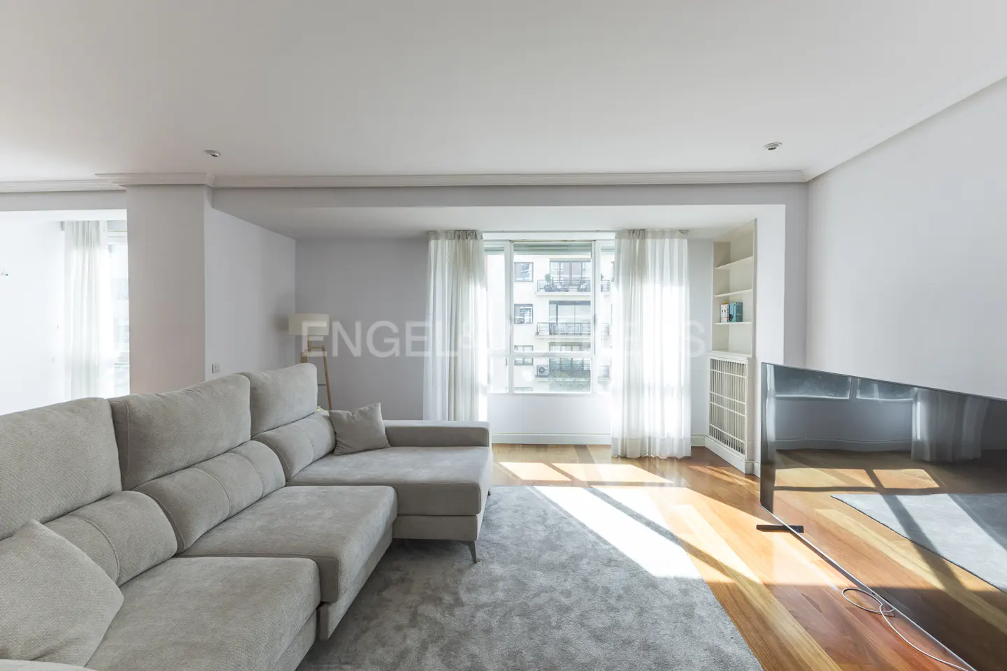 Bright living room with a large gray sectional sofa, wood floors, and a large window with sheer white curtains. A gray rug sits in the center.